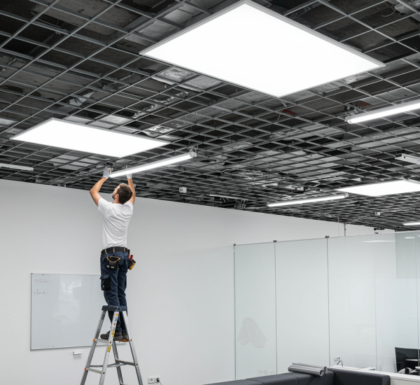 "Instalador ajustando un panel de techo suspendido en una oficina moderna con iluminación LED, paredes blancas y mobiliario contemporáneo."